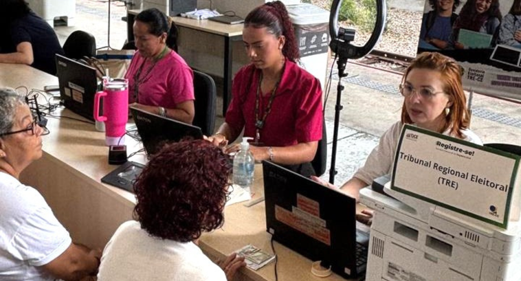 Imagem mostra um balcão de atendimento com quatro pessoas sentadas atrás de computadores e duas ...
