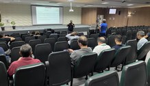 A mostra o auditório com fileiras de cadeiras, algumas ocupadas por pessoas sentadas, voltadas p...