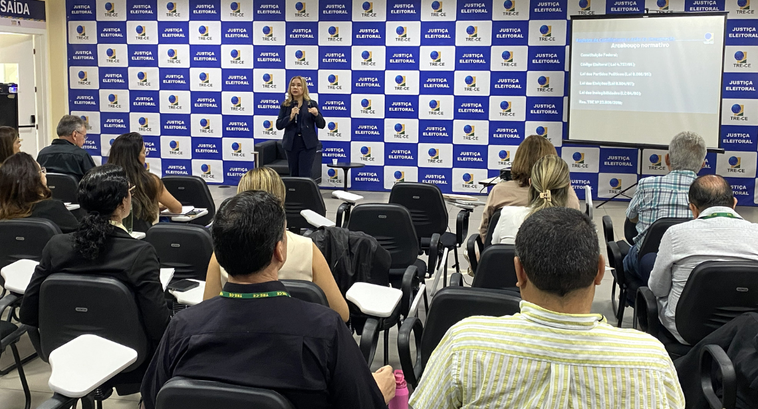 Na imagem, mostra em primeiro plano, fileiras de cadeiras ocupadas por participantes voltados pa...