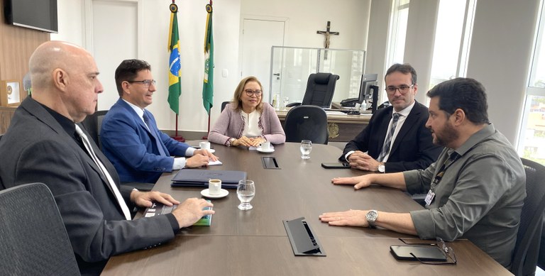 Na imagem, na mesma sala, as cinco pessoas estão sentadas ao redor de uma mesa retangular. Sobre...