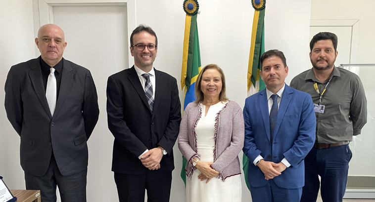 Na imagem, cinco pessoas estão em pé lado a lado, posicionadas em frente de duas bandeiras, uma ...