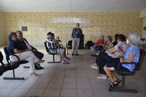 A imagem mostra um grupo de pessoas sentadas em cadeiras distribuídas pelo espaço, entre homens ...