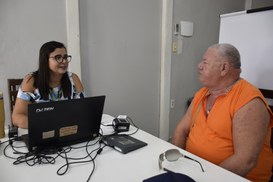 A imagem mostra uma mulher, à esquerda, e um homem, à direita, sentados ao redor de uma mesa bra...