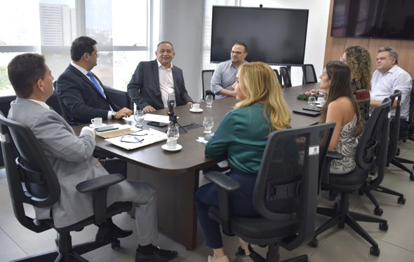 Na imagem, as pessoas aparecem reunidas ao redor de uma mesa de reuniões. Todas estão sentadas e...