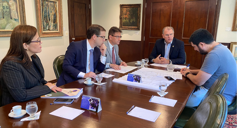 A imagem mostra uma reunião com cinco pessoas sentadas ao redor de uma mesa retangular de madeir...
