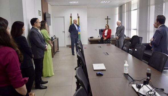 A imagem mostra, de forma mais ampla, a sala e os participantes da cerimônia. Na foto, há nove p...