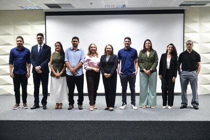 A imagem mostra dez pessoas alinhadas em pé, lado a lado, sobre o palco do auditório do TRE-CE. ...