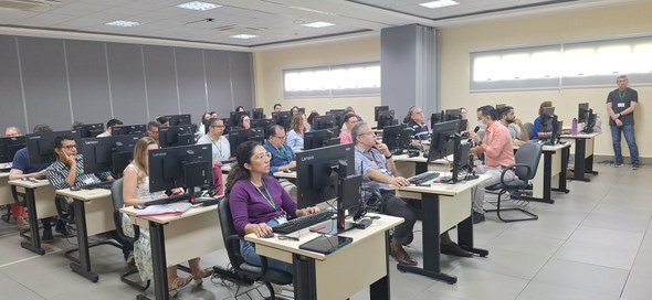 A imagem mostra, de outro ângulo, os participantes da reunião de frente, sentados ao redor da me...