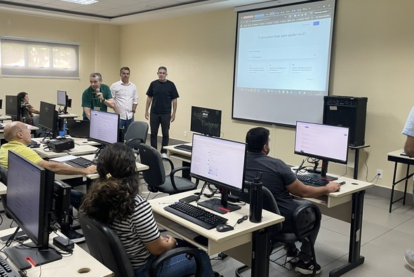 A imagem mostra, em primeiro ângulo, os participantes diante dos computadores e ao fundo, três h...