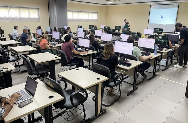 A imagem mostra a sala de treinamento da sede do TRE-CE com várias fileiras de mesas e cadeiras ...