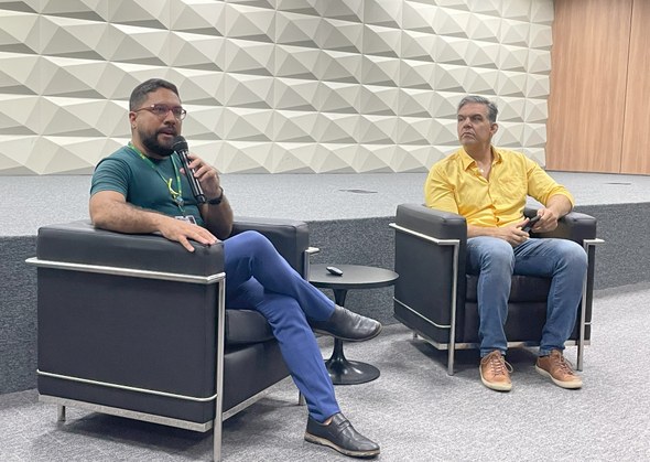 No registro, dois homens estão sentados em poltronas pretas. Um deles, à esquerda, segura o micr...