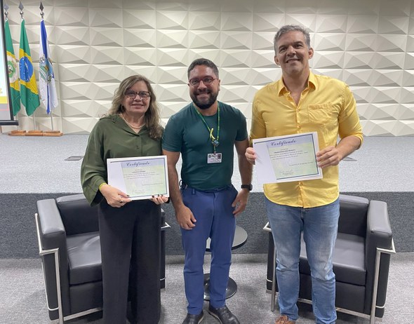 A foto apresenta três pessoas — dois homens e uma mulher — posando lado a lado. Os dois que estã...
