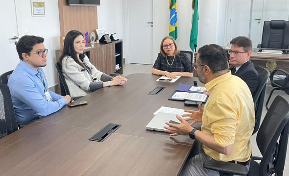 Na imagem, na mesma sala, as cinco pessoas estão sentadas ao redor de uma mesa retangular. Ao fu...