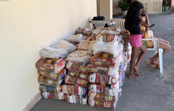 Na imagem, há diversos kits de alimentos organizados em pilhas sobre o piso, encostados em uma p...