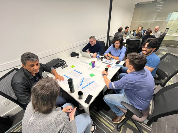 A imagem mostra um grupo de pessoas em um ambiente externo, sorrindo para a foto, com uma divisó...