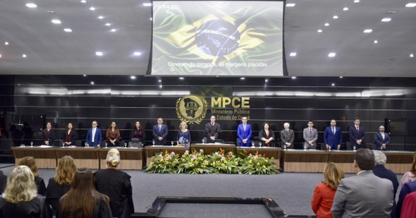 A imagem mostra a mesa de honra do evento. Sobre o palco, há uma grande mesa retangular de madei...