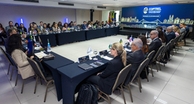 Em uma sala de eventos, dezenas de pessoas estão sentadas ao redor de mesas organizadas em forma...