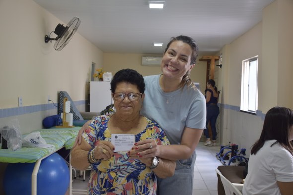 Na imagem, uma mulher idosa está em pé segurando um documento diante da câmera, acompanhada pela...