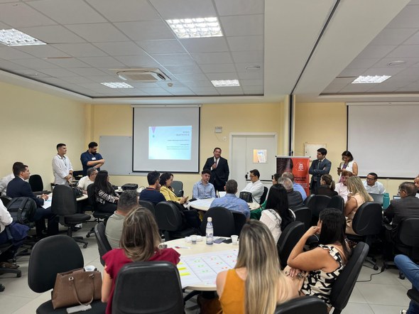 Registro fotográfico com diversas pessoas sentadas diante de mesas brancas circulares. Em pé, co...