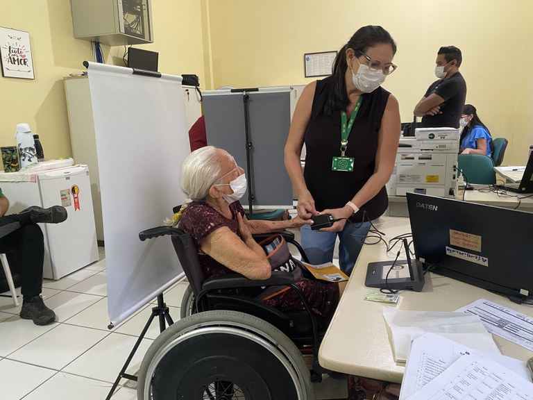 Na imagem, uma colaboradora do TRE-CE está em pé ao lado de uma mesa e segura o dedo indicador d...