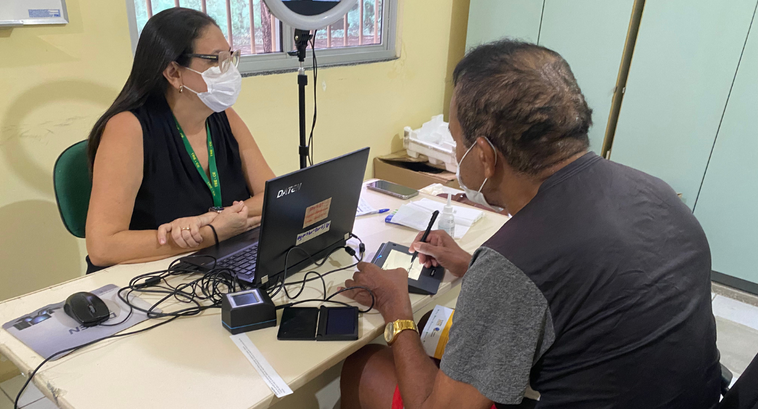 Na imagem, um homem idoso está sentado numa cadeira de rodas em frente a uma colaboradora do TRE...
