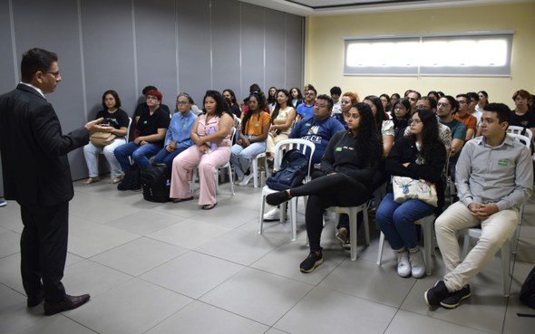 Na imagem, os estudantes estão sentados em cadeiras brancas dentro de uma sala fechada. O juiz a...
