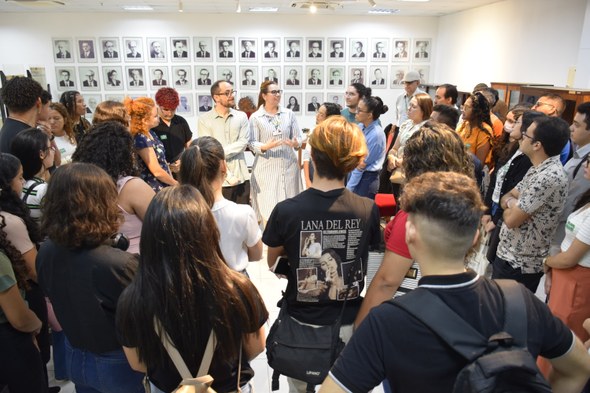 Na imagem, o grupo está em uma sala ampla, em pé, formando um círculo ao redor de duas pessoas q...