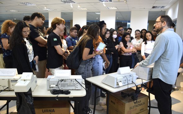 Na imagem, os jovens estão em pé ao redor de mesas que exibem modelos antigos de urnas eletrônic...