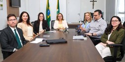 A imagem mostra uma reunião em uma sala, com sete pessoas sentadas ao redor de uma mesa retangul...
