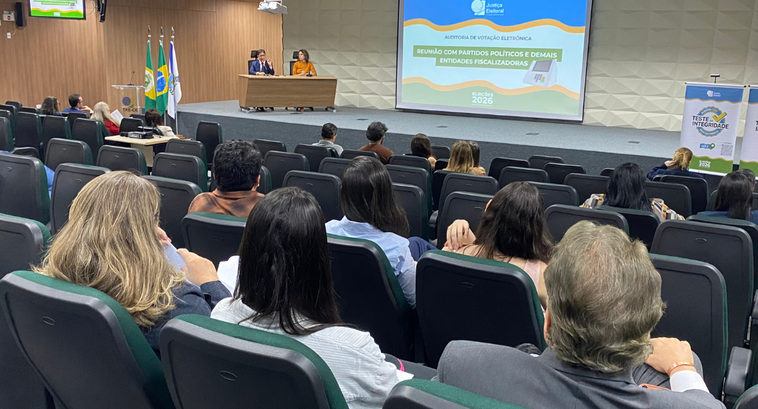 A imagem mostra um auditório com pessoas sentadas em fileiras de cadeiras, voltadas para um palc...