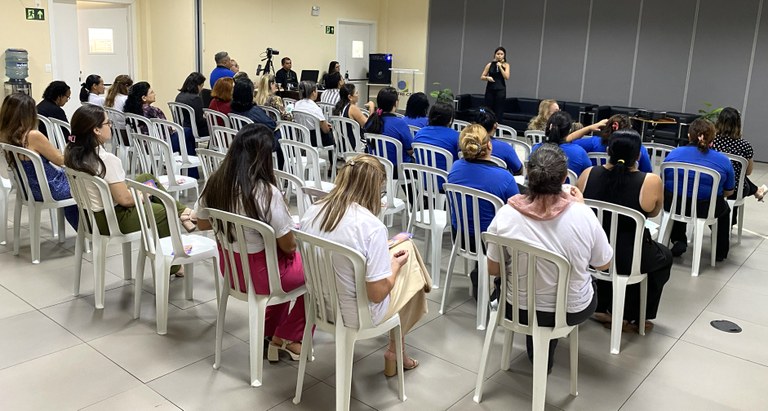 O ambiente mostra um público de costas sentado em cadeiras de plásticos, na sala de treinamento ...