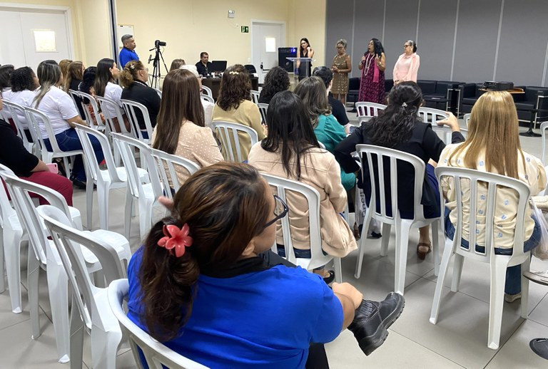 O ambiente mostra um público de costas sentado em cadeiras de plásticos, na sala de treinamento ...