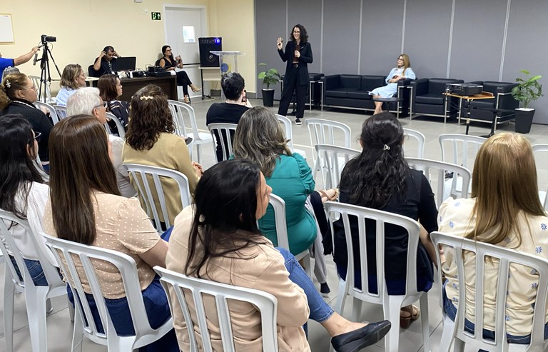 O ambiente mostra um público de costas sentado em cadeiras de plásticos, na sala de treinamento ...