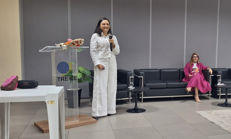 A imagem, no ambiente de uma sala de treinamento do TRE-CE, com parede cinza. No local há um púl...