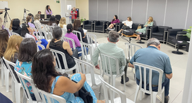 O ambiente mostra um público de costas sentado em cadeiras de plásticos, na sala de treinamento ...