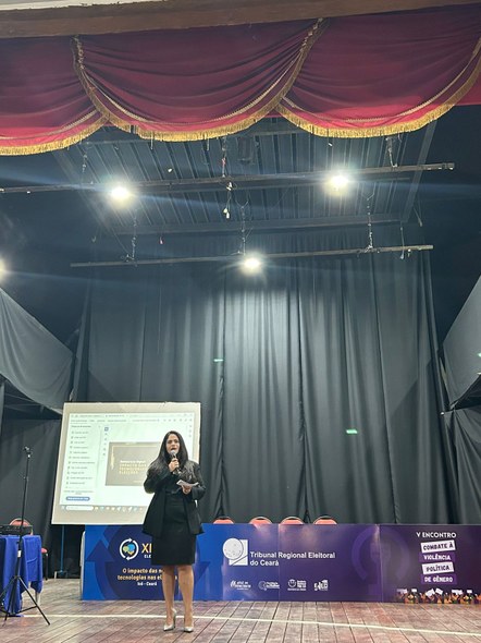 Na foto, uma mulher veste preto e está sobre um palco segurando um microfone. Atrás dela, há a m...