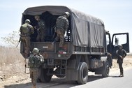 A imagem mostra um caminhão militar verde com cobertura de lona cinza estacionado em uma via asf...
