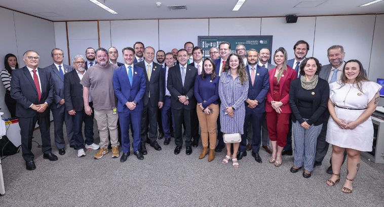 A imagem é um registro de um grupo de pessoas reunidas em uma sala. Ao todo, cerca de 25 pessoas...