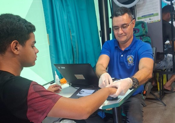A imagem mostra dois homens sentados frente a frente em uma mesa, durante um atendimento eleitor...