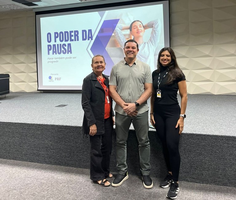 Na imagem, três pessoas estão em pé sobre um palco, lado a lado, voltadas para a câmera. Atrás d...